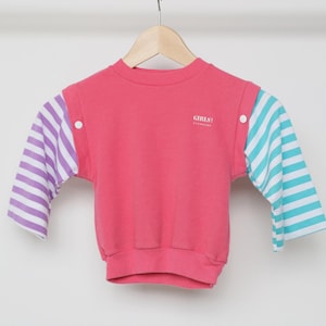 May include: A pink sweatshirt with striped sleeves in purple and white on one side and turquoise and white on the other. The text "GIRLS" is printed on the chest. The garment is hanging on a wooden hanger.
