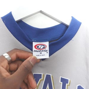 Puede incluir: Una camiseta gris con cuello y ribetes azules, colgada de una percha blanca. La camiseta tiene una etiqueta que dice "TRUEFAN" y "MADE IN HAITI". La camiseta es talla M.