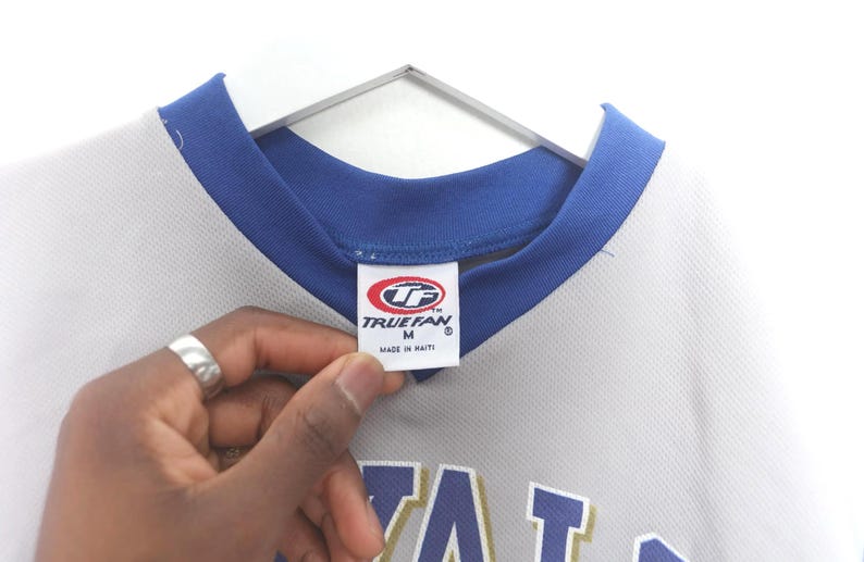 Puede incluir: Una camiseta gris con cuello y ribetes azules, colgada de una percha blanca. La camiseta tiene una etiqueta que dice "TRUEFAN" y "MADE IN HAITI". La camiseta es talla M.