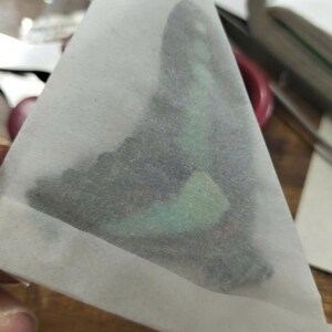 K&ouml;nnte beinhalten: Ein dreieckiges St&uuml;ck halbtransparentes wei&szlig;es Papier, das einen dunklen Schmetterlingsfl&uuml;gel-Aufn&auml;her bedeckt. Der Fl&uuml;gel zeigt ein markantes, schillerndes mintgr&uuml;nes Band und subtile schwarze Muster, die durch das d&uuml;nne Schutzpapier sichtbar sind.
