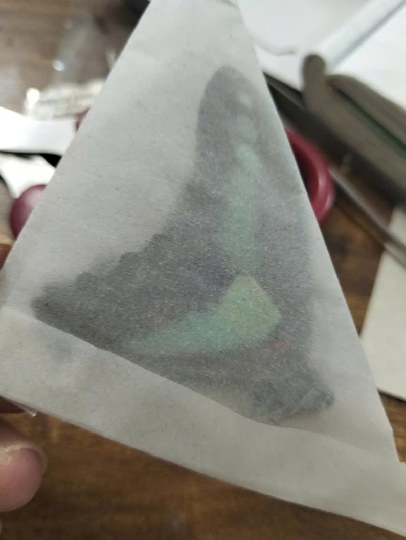 K&ouml;nnte beinhalten: Ein dreieckiges St&uuml;ck halbtransparentes wei&szlig;es Papier, das einen dunklen Schmetterlingsfl&uuml;gel-Aufn&auml;her bedeckt. Der Fl&uuml;gel zeigt ein markantes, schillerndes mintgr&uuml;nes Band und subtile schwarze Muster, die durch das d&uuml;nne Schutzpapier sichtbar sind.