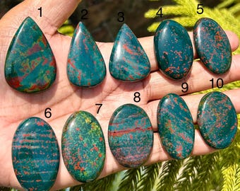 May include: A collection of ten polished bloodstone gemstones in various shapes, including teardrop and oval. The stones display a deep green color with red speckles and veins, showcasing natural patterns.