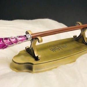 Puede incluir: Una varita decorativa con un mango rosa y un eje marrón, apoyada en un soporte dorado. El soporte está inscrito con "Harry Potter". La varita es un artículo de colección.