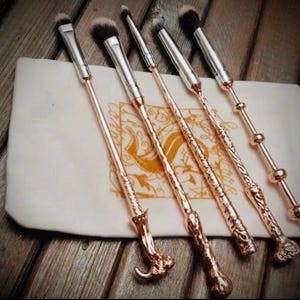 Pu&ograve; includere: Un set di cinque pennelli per trucco color oro rosa con intricati manici a forma di bacchetta, disposti su una pochette color crema. I pennelli hanno testine argentate e sono esposti su una superficie di legno.