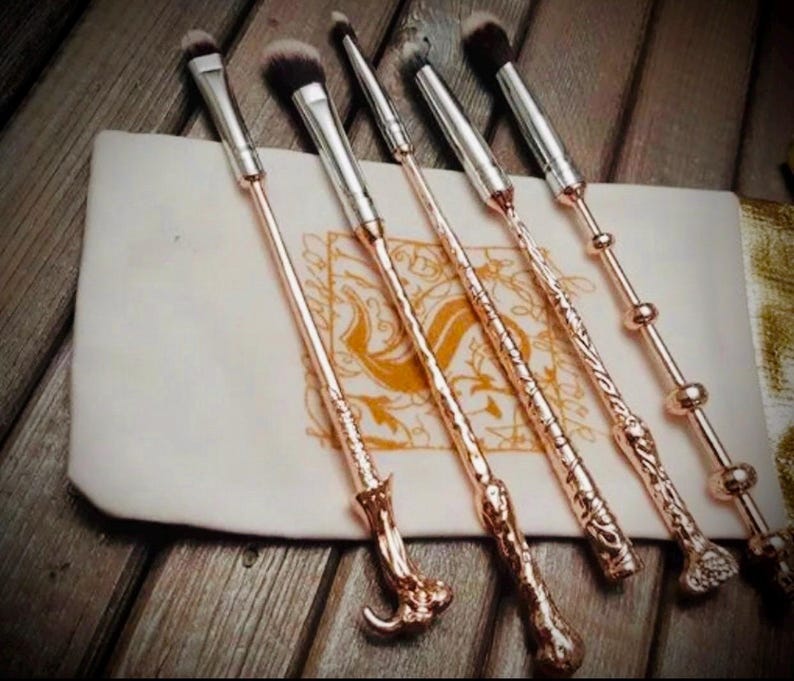 Pu&ograve; includere: Un set di cinque pennelli per trucco color oro rosa con intricati manici a forma di bacchetta, disposti su una pochette color crema. I pennelli hanno testine argentate e sono esposti su una superficie di legno.