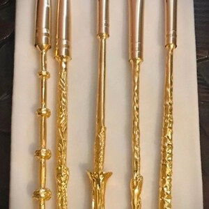 Pu&ograve; includere: Un set di sei pennelli per il trucco con manici color oro e setole scure. I manici presentano disegni decorati, tra cui motivi floreali e geometrici. I pennelli sono disposti su una superficie bianca.