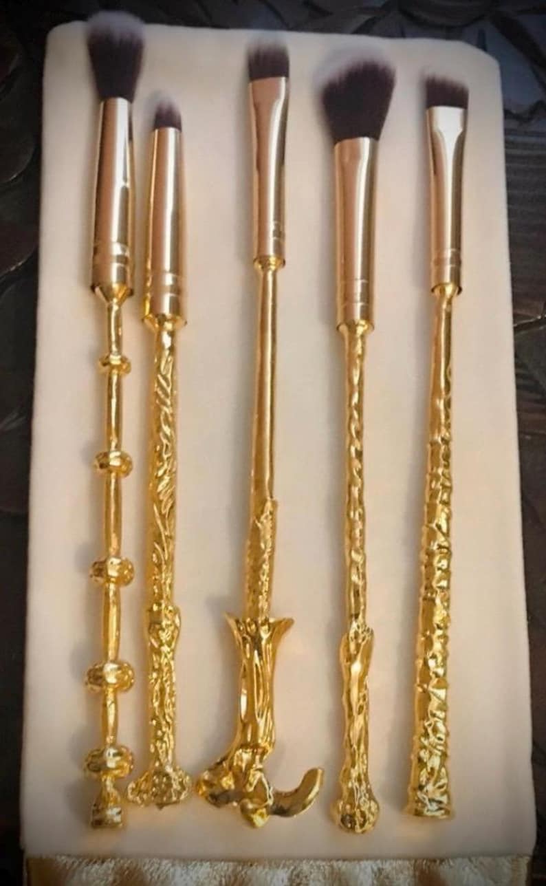 Pu&ograve; includere: Un set di sei pennelli per il trucco con manici color oro e setole scure. I manici presentano disegni decorati, tra cui motivi floreali e geometrici. I pennelli sono disposti su una superficie bianca.