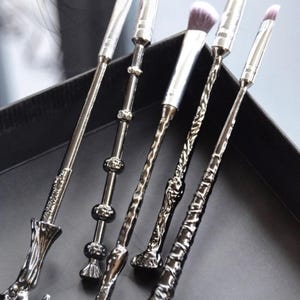 May include: A set of six silver-toned makeup brushes with ornate wand-like handles. The brushes have white and purple bristles and are arranged in a black box. The handles feature various decorative designs, including spheres and textured patterns.