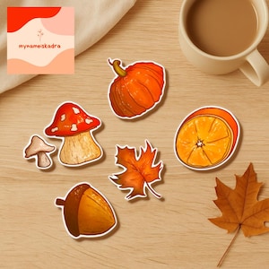 Könnte beinhalten: Eine Sammlung von Aufklebern zum Thema Herbst, darunter ein Kürbis, ein Pilz, eine Eichel, eine Orangenscheibe und ein Ahornblatt. Die Aufkleber sind in Orange-, Braun- und Beigetönen gehalten. Der Text "mynameiskadra" ist sichtbar.