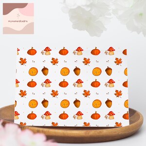 Könnte beinhalten: Eine weiße Karte mit einem sich wiederholenden Muster aus orangefarbenen Kürbissen, Pilzen, Ahornblättern, Eicheln und Orangenscheiben. Die Karte liegt auf einem Holztablett. Der Text "mynameiskadra" befindet sich oben links.