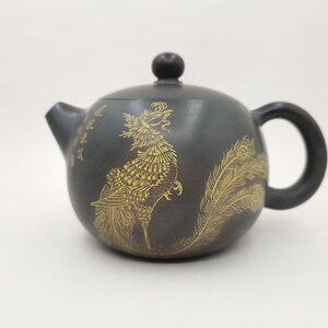 Könnte beinhalten: Dunkelgraue Keramik-Teekanne mit goldenem Phönix-Design. Die Teekanne hat einen abgerundeten Körper, einen gebogenen Ausguss und einen Schlaufengriff. Der Deckel hat einen kleinen, runden Knopf. Der Phönix ist mit detaillierten Federn dargestellt.
