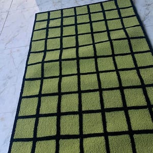 K&ouml;nnte beinhalten: Ein rechteckiger Teppich mit einem Gittermuster aus hellgr&uuml;nen Quadraten, die durch dicke schwarze Linien getrennt sind. Der Teppich hat eine strukturierte, getuftete Oberfl&auml;che und einen durchgehenden schwarzen Rand um das Karomuster.