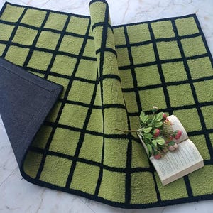 K&ouml;nnte beinhalten: Ein rechteckiger Teppich mit einer lindgr&uuml;nen Oberfl&auml;che und einem schwarzen Gittermuster. Der Teppich ist teilweise gefaltet und gibt den Blick auf eine einfarbig schwarze Unterseite frei. Ein kleiner Strau&szlig; rosa und wei&szlig;er Kunstblumen liegt auf einem offenen Buch.