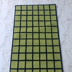 K&ouml;nnte beinhalten: Ein rechteckiger Teppich mit einem Gittermuster aus hellgr&uuml;nen Quadraten, die durch d&uuml;nne schwarze Linien getrennt sind. Der Teppich hat eine strukturierte Oberfl&auml;che und ist von einem durchgehenden schwarzen Rand umgeben.