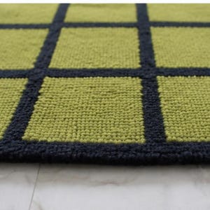 K&ouml;nnte beinhalten: Ein rechteckiger Teppich mit einem Gittermuster aus lindgr&uuml;nen Quadraten, die durch dicke, dunkelmarineblaue Linien getrennt sind. Der Teppich hat eine strukturierte, getuftete Oberfl&auml;che mit einer gleichm&auml;&szlig;igen Florh&ouml;he &uuml;ber die gesamte Fl&auml;che.