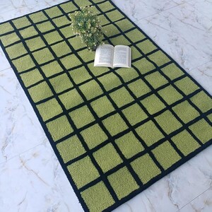 K&ouml;nnte beinhalten: Ein rechteckiger Teppich mit einem Gittermuster aus hellgr&uuml;nen Quadraten, die durch dicke schwarze Linien getrennt sind. Der Teppich hat eine strukturierte, getuftete Oberfl&auml;che. Eine kleine Pflanze mit wei&szlig;en Bl&uuml;ten und ein offenes Buch liegen darauf.