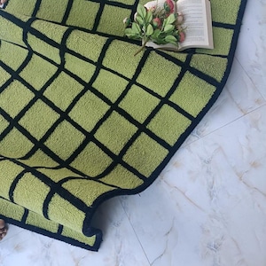 Può includere: Un tappeto rettangolare con un motivo a griglia di quadrati verde lime delineati da spesse linee nere. Un libro aperto con pagine bianche e un piccolo bouquet di fiori artificiali rosa e verdi sono posizionati sulla superficie del tappeto.