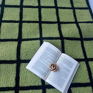 K&ouml;nnte beinhalten: Ein strukturierter Teppich mit einem Gittermuster aus hellgr&uuml;nen Quadraten, die durch dicke schwarze Linien getrennt sind. Ein offenes Buch mit wei&szlig;en Seiten und schwarzem Text liegt flach auf dem Teppich, mit einer kleinen hellbraunen Kunstblume.