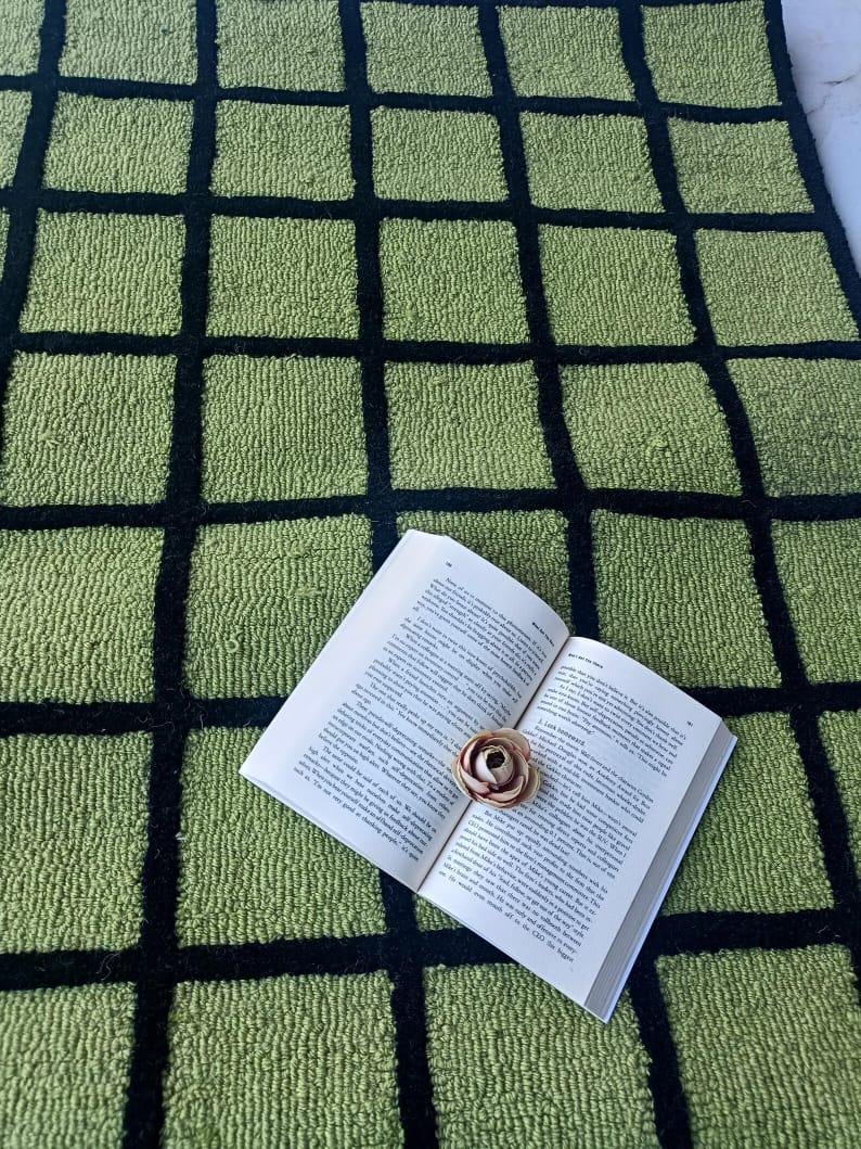 K&ouml;nnte beinhalten: Ein strukturierter Teppich mit einem Gittermuster aus hellgr&uuml;nen Quadraten, die durch dicke schwarze Linien getrennt sind. Ein offenes Buch mit wei&szlig;en Seiten und schwarzem Text liegt flach auf dem Teppich, mit einer kleinen hellbraunen Kunstblume.