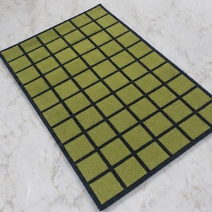 K&ouml;nnte beinhalten: Ein rechteckiger Teppich mit einem Gittermuster aus olivgr&uuml;nen Quadraten, die durch d&uuml;nne schwarze Linien getrennt sind. Der Teppich hat einen durchgehenden schwarzen Rand, der das gesamte Design einrahmt und ein strukturiertes, geometrisches Aussehen erzeugt.