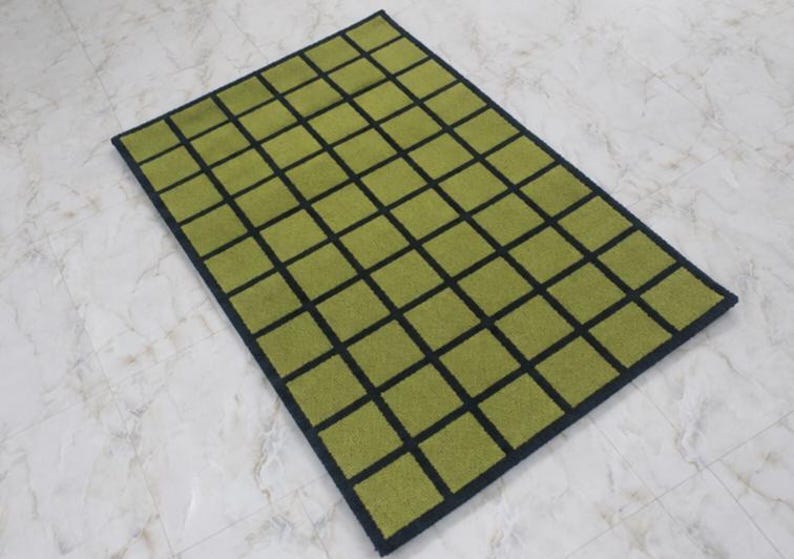 K&ouml;nnte beinhalten: Ein rechteckiger Teppich mit einem Gittermuster aus olivgr&uuml;nen Quadraten, die durch d&uuml;nne schwarze Linien getrennt sind. Der Teppich hat einen durchgehenden schwarzen Rand, der das gesamte Design einrahmt und ein strukturiertes, geometrisches Aussehen erzeugt.