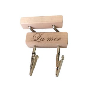May include: A wooden memo holder with two metal clips. The holder has two wooden blocks, one above the other, connected by metal loops. The bottom block is engraved with the words "La mer".