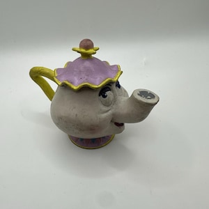 May include: A ceramic teapot with a cartoon face, featuring a light gray body, a purple lid with yellow trim, and a yellow handle. The teapot has a smiling expression and is set against a white background.