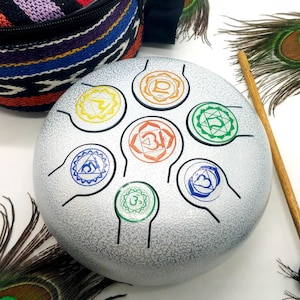May include: A silver tongue drum with colorful chakra symbols and a wooden mallet. A patterned carrying case with a zipper is also present. Peacock feathers are arranged around the drum and case.