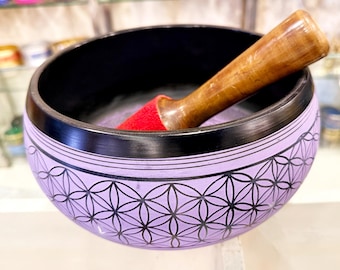 May include: A lavender singing bowl with a black rim and a geometric pattern. A wooden mallet rests inside the bowl on a red cushion. The bowl is designed for meditation and sound healing.