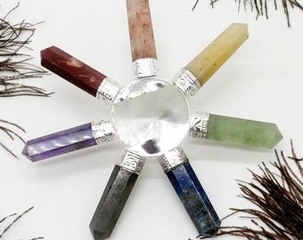 Può includere: Un generatore di energia dei chakra con sette punte di cristallo colorate che irradiano da un disco centrale argentato. I cristalli sono di vari colori, tra cui rosso, viola, verde e blu. Su uno sfondo bianco con piume di pavone.