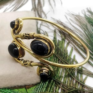 Peut inclure: Un bracelet en métal doré orné de quatre pierres précieuses noires ovales. Le bracelet présente un design ouvert unique avec des volutes décoratives et des accents enroulés. Le fond comprend des plumes de paon vertes et brunes.