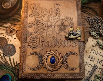 May include: A tan leather journal with the embossed text Book Of Shadows, a pentagram, and triple moon symbols. The cover features a central blue gemstone set in a woven leather frame and a brass-colored metal latch closure on the right side.