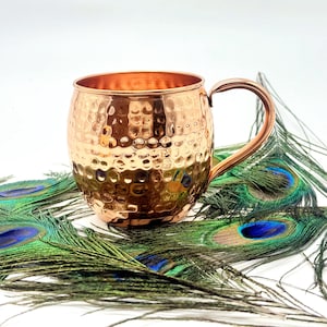 Könnte beinhalten: Ein kupferner Moscow Mule-Becher mit gehämmertem Muster und einem gebogenen Griff. Der Becher wird mit mehreren grünen und blauen Pfauenfedern gezeigt.