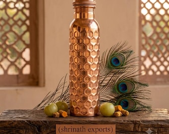 Op de afbeelding: Een cilindrische koperen waterfles met een verhoogd zeshoekig honingraatpatroon op het buitenoppervlak. De fles heeft een metallic afwerking en een schroefdop. Een rechthoekig plaatje aan de basis vermeldt (shrinath exports) FINE ARTISANAL COPPER MADE IN JAIPUR.