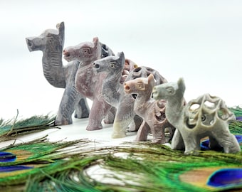 May include: A collection of carved stone camel figurines in varying shades of gray and pink. The camels are arranged in a row, with intricate details. The figurines are displayed on a bed of green and blue peacock feathers.