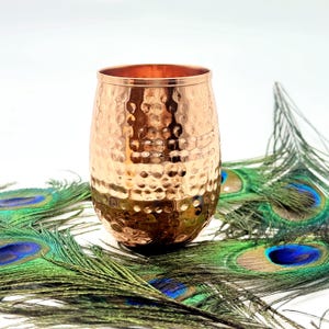 Puede incluir: Un vaso de color cobre con acabado martillado. La copa tiene forma redondeada y una abertura ancha. El fondo presenta varias plumas de pavo real con tonos verdes, azules y marrones. El vaso se utiliza probablemente para bebidas.