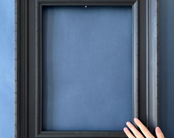 May include: A black picture frame with a rectangular opening. The frame has a detailed, textured design with multiple layers and a dark finish. The frame is displayed against a blue background.