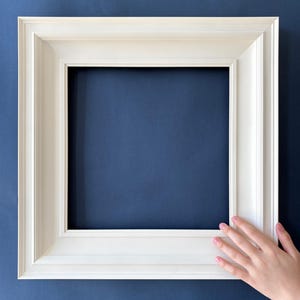 Pode incluir: Uma moldura quadrada, creme, com um design detalhado e em camadas. A moldura está sobre um fundo azul escuro. A moldura está vazia, pronta para uma foto ou obra de arte.