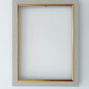 May include: A rectangular picture frame featuring a wide, light gray fabric-textured border surrounding a thin, metallic gold inner trim. The frame is empty, showcasing a clean, structured design with a neutral color palette and a classic finish.