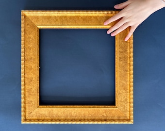 May include: A square, ornate gold picture frame with a textured surface. The frame is held by a hand with pink nail polish against a dark blue background. The frame's interior is empty.
