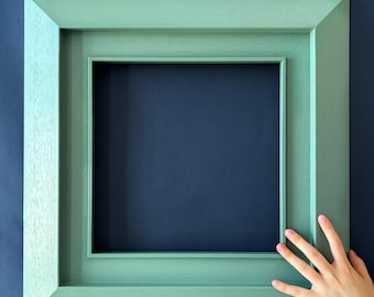 May include: A square, pale green picture frame against a dark blue backdrop. The frame has a layered design and an empty centre. A hand with a silver bracelet is touching the frame.