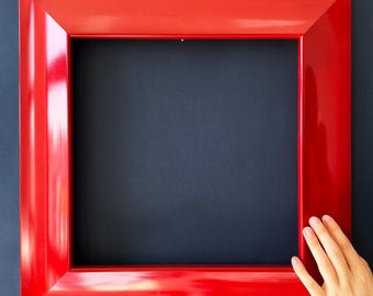 May include: A square, glossy red picture frame. The frame is a vibrant, solid red colour and has a thick, bevelled edge. The frame is empty, with a dark blue background. A hand is touching the frame.