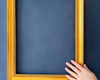 May include: A rectangular, ornate gold picture frame with a detailed beaded inner edge. The frame is empty, with a dark blue background. A hand is partially visible, touching the frame.