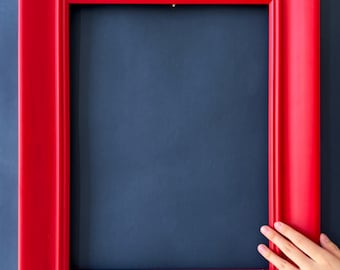 May include: A bright red picture frame with a rectangular design. The frame is set against a dark blue background. A hand is touching the frame.
