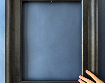 May include: A rectangular black picture frame with a grooved texture. The frame is empty, with a dark blue background visible through the opening. The frame is held by a hand with pink nail polish.