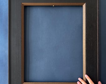 May include: A rectangular black picture frame with a brown inner border. The frame is empty, with a dark blue background visible through the opening. The frame is held by a hand.