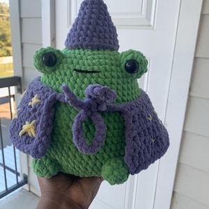 May include: A handmade green crochet frog wearing a purple wizard hat and cape, adorned with yellow stars. The frog has large black eyes and a yellow star on top of its hat. The cape is tied with a purple cord.