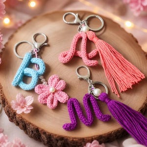 May include: Three handmade crochet letter keychains in light blue, pink, and purple. The blue letter B features a pink crochet flower, while the pink letter A and purple letter M each include a matching long yarn tassel and a single white pearl bead.