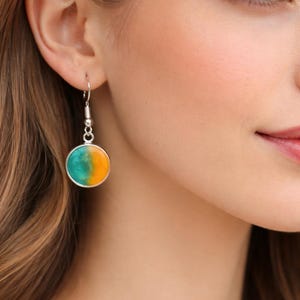 May include: A silver earring with a circular pendant. The pendant is divided into two colours: teal and orange. The earring is hanging from the ear of a person with fair skin and brown hair.