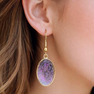 May include: Gold-tone drop earrings featuring an oval pendant filled with purple and silver glitter. The earrings have a hook fastening and a small gold bead. The earrings are shown on a person.